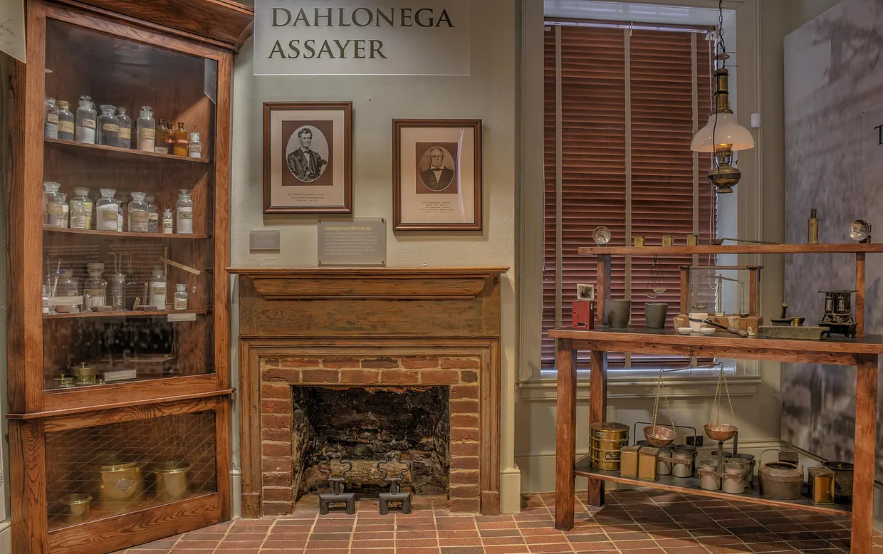 A museum room set up as an assay office: a glass-fronted cabinet of bottles, two framed portraits above a brick fireplace under a sign reading Dahlonega Assayer, and a wooden worktable of scales and tins by a window.