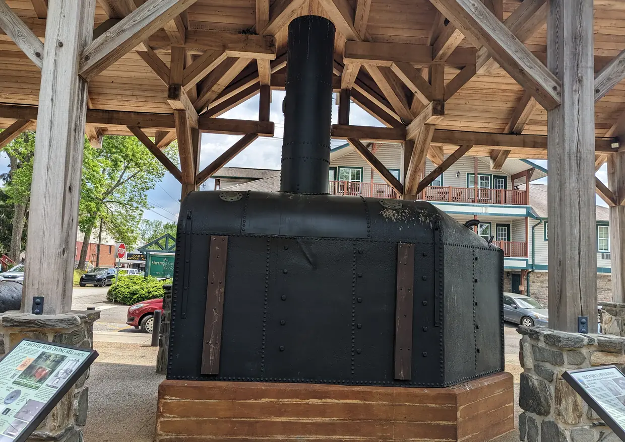 A large riveted black iron chamber with a tall stack on a timber base, under an open wooden shelter with stone piers and interpretive signs, a building with balconies and parked cars behind.