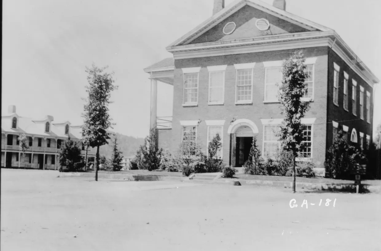 A black-and-white photograph of a two-storey brick courthouse with a pedimented gable, two round attic windows and a side portico, young trees on a bare dirt square and a long galleried building at the far left.