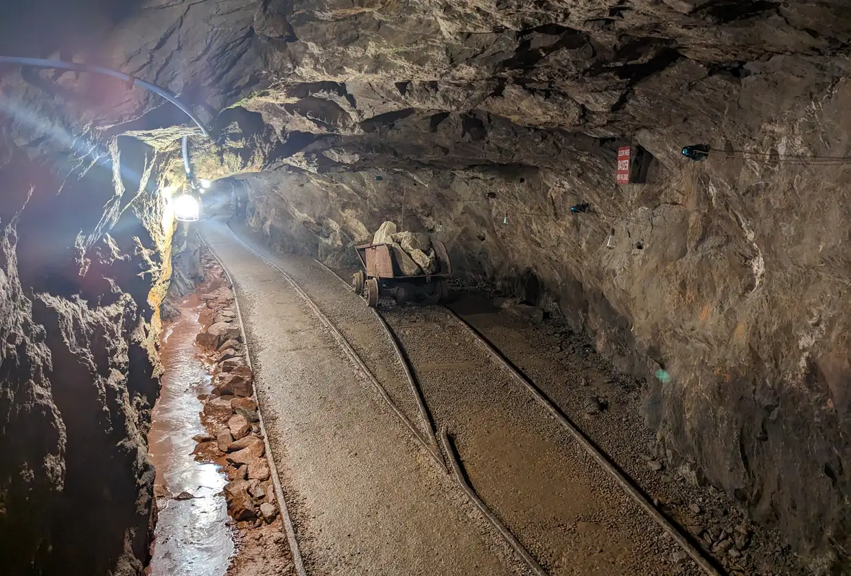 A narrow rock tunnel lit by a lamp, with a small ore cart standing on rails and a shallow channel of water along the left wall.