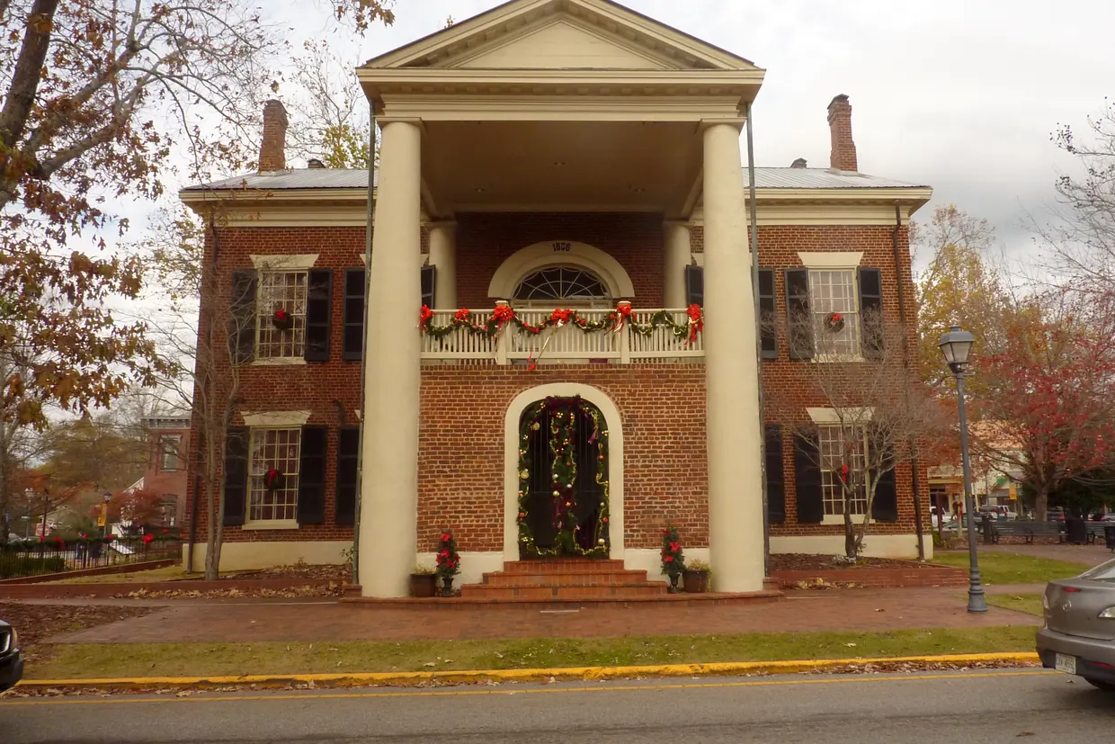 The red-brick museum front with tall white columns, its arched doorway, balcony rail and windows hung with green wreaths and red bows, autumn trees on either side and a lamp post at the curb.
