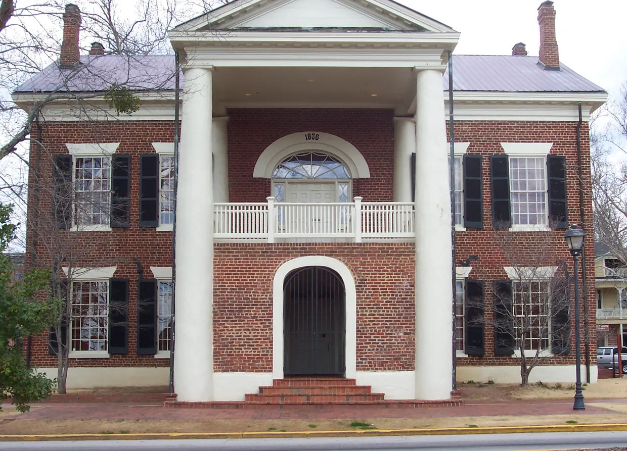 A two-storey red-brick building with a white-columned portico, a fanlit balcony door above an arched, gated entrance, black shutters and two end chimneys, seen straight on across a brick walk in winter.