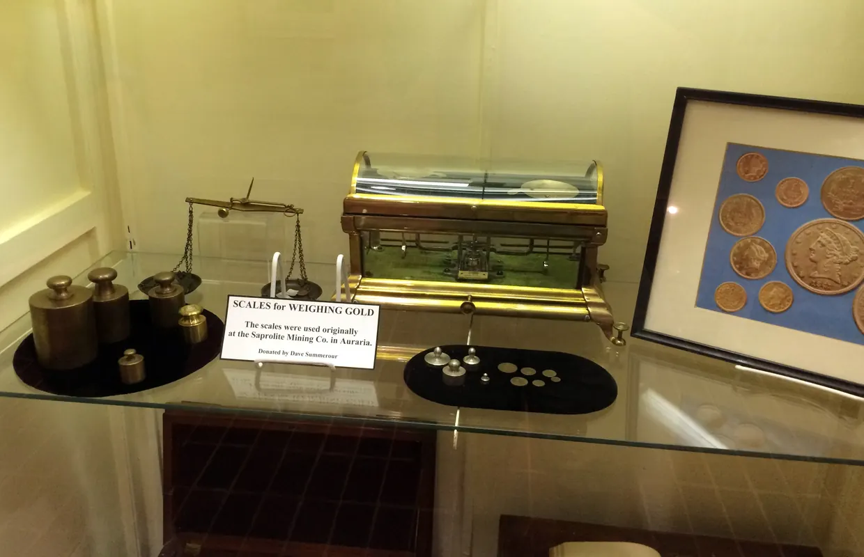 A museum case of brass weights and balances beside a card reading Scales for weighing gold, used originally at the Saprolite Mining Co. in Auraria, with a framed display of coins.