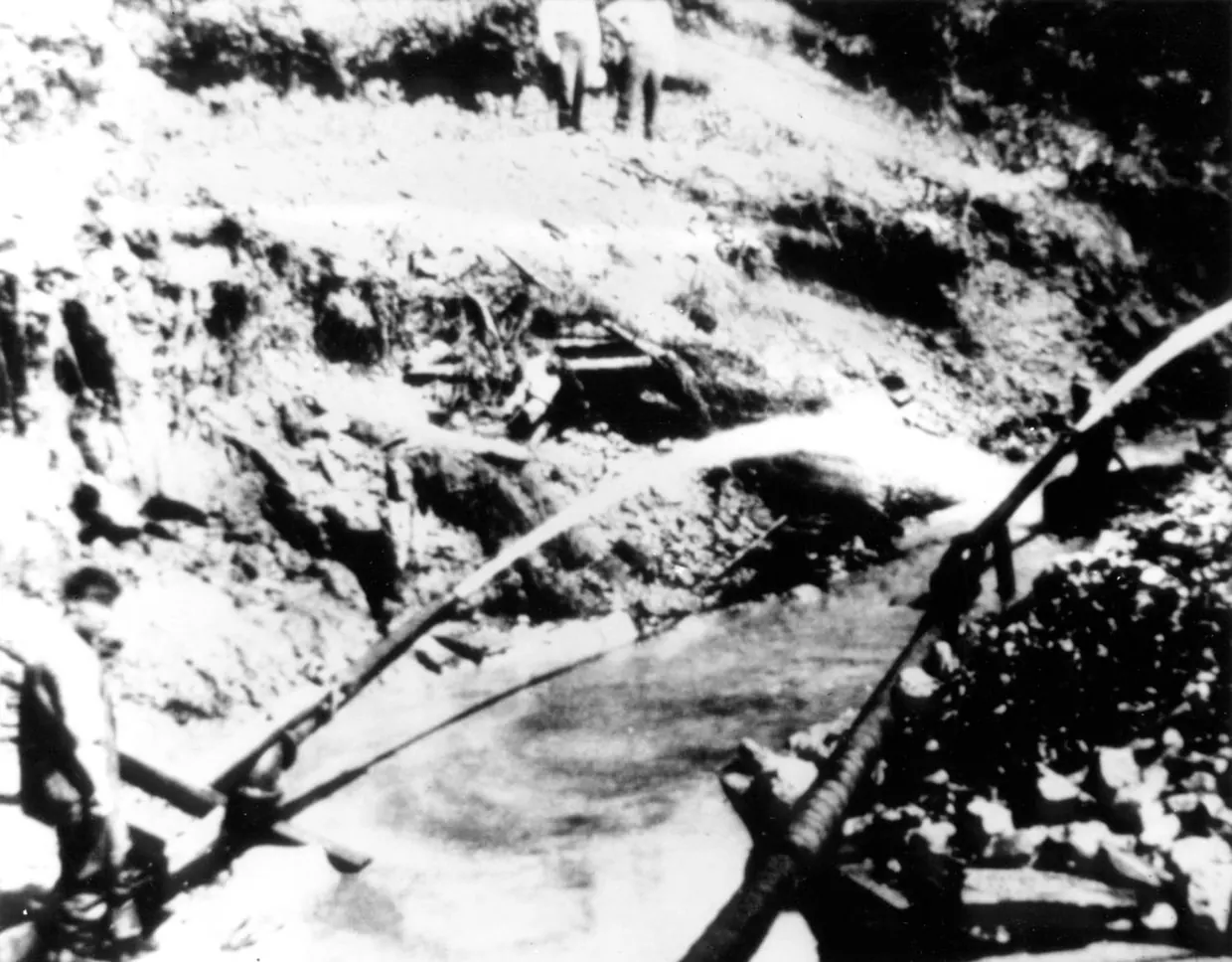 A grainy black-and-white photograph of a raw, washed-out hillside above a muddy pool, a long pale arc of water crossing it, timber pipes in the foreground and men standing on the bank.