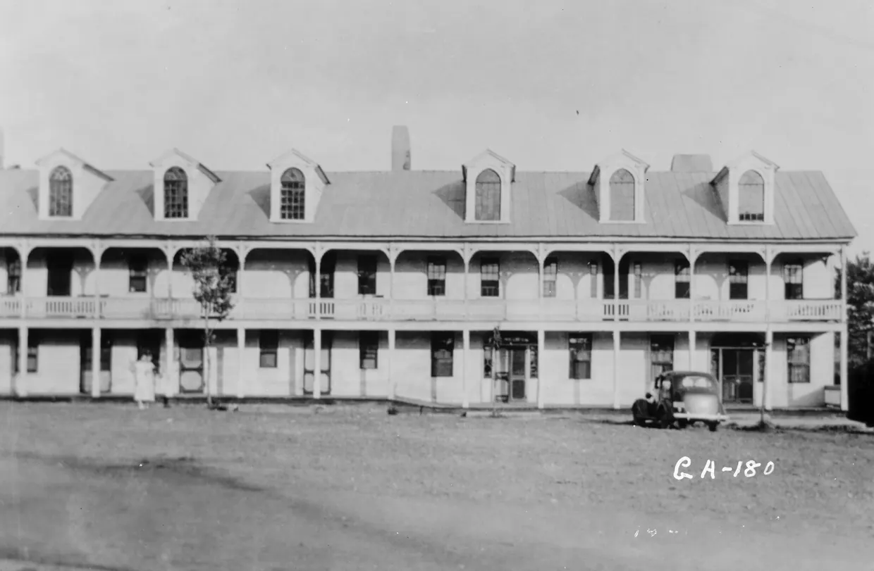 A black-and-white photograph of a long two-storey building with a row of dormers in its metal roof and full-length porches on both floors, an old car parked at the right on a bare dirt square.