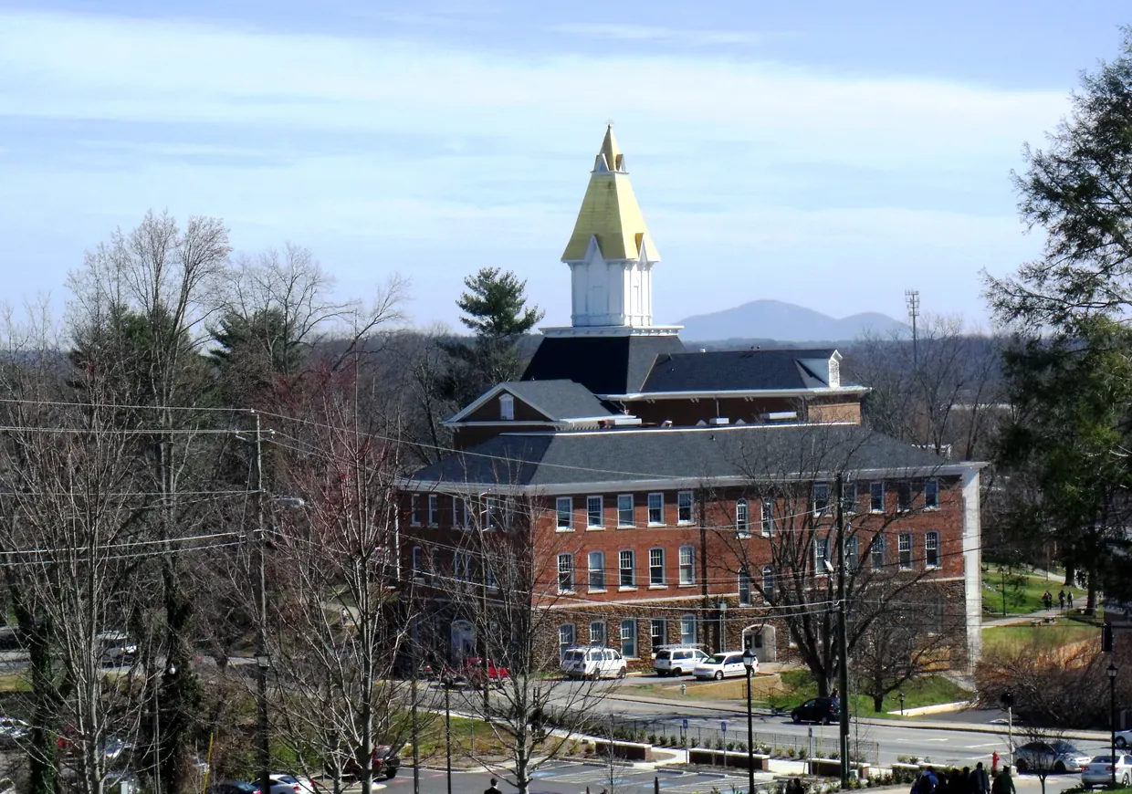 A gold-coloured steeple rises above a red-brick hall with a dark mansard roof, seen across a campus street and lawn with bare early-spring trees, a blue mountain on the horizon.