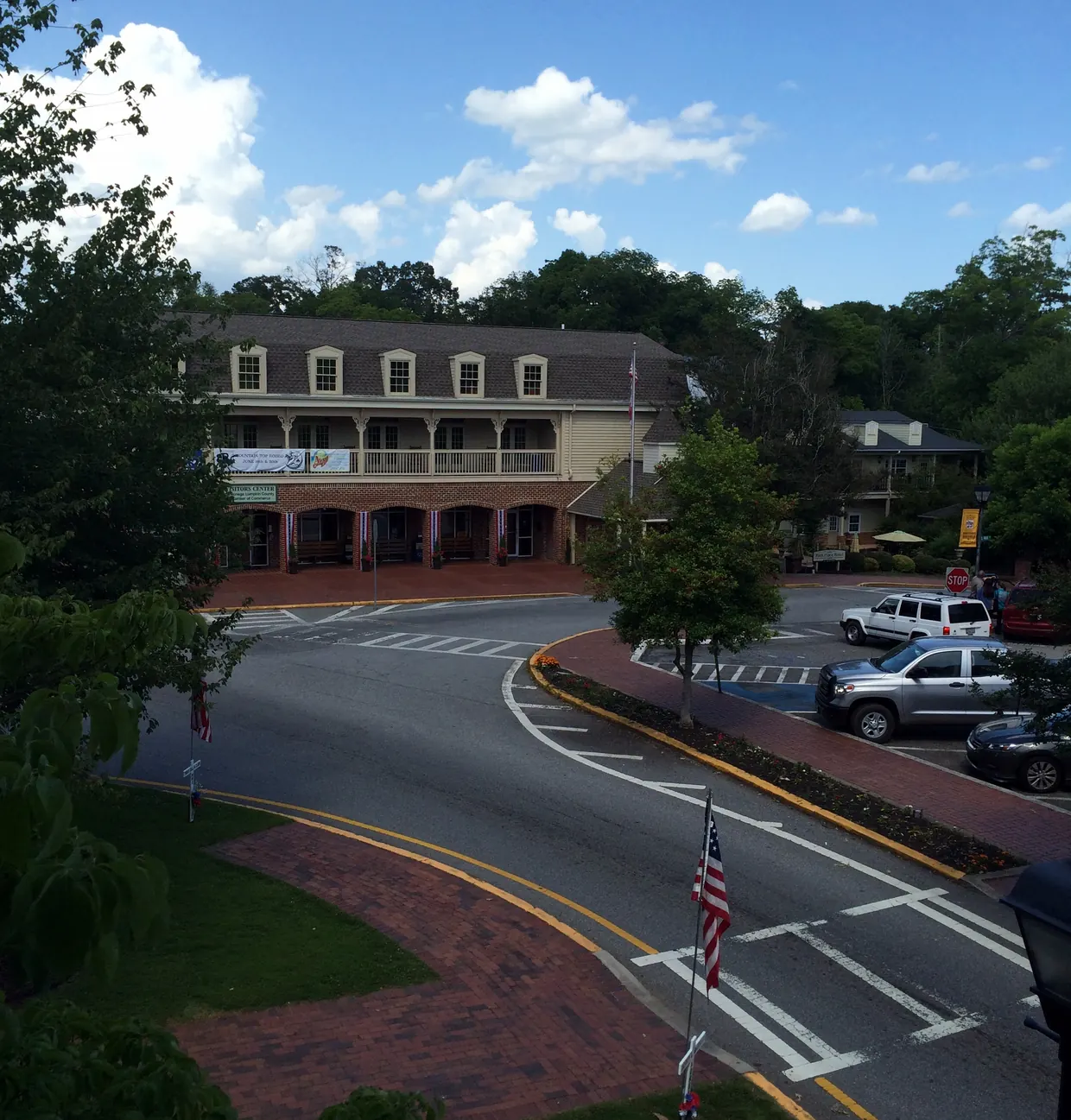 A long two-storey building with a row of dormer windows and a full-length upper porch faces a curving street, parked cars and a brick-edged lawn with an American flag, with wooded hills behind.