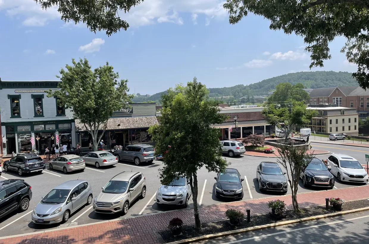 A high view over a parking lot full of cars to a row of low shopfronts under shade trees, with a brick building and a green wooded hill beyond under a blue sky.