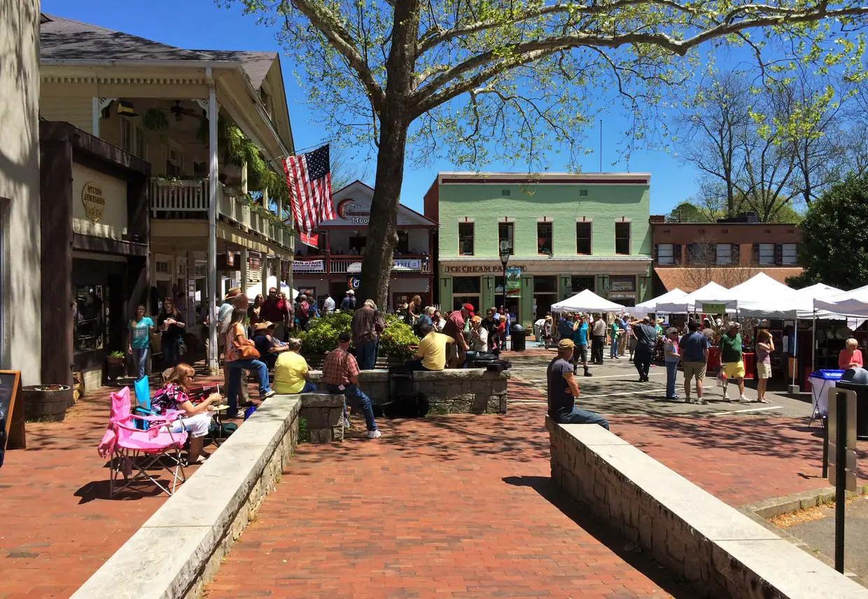 A brick sidewalk under a large shade tree busy with people, white vendor tents along the far side, a covered porch with an American flag at left and a pale-green two-storey storefront behind.