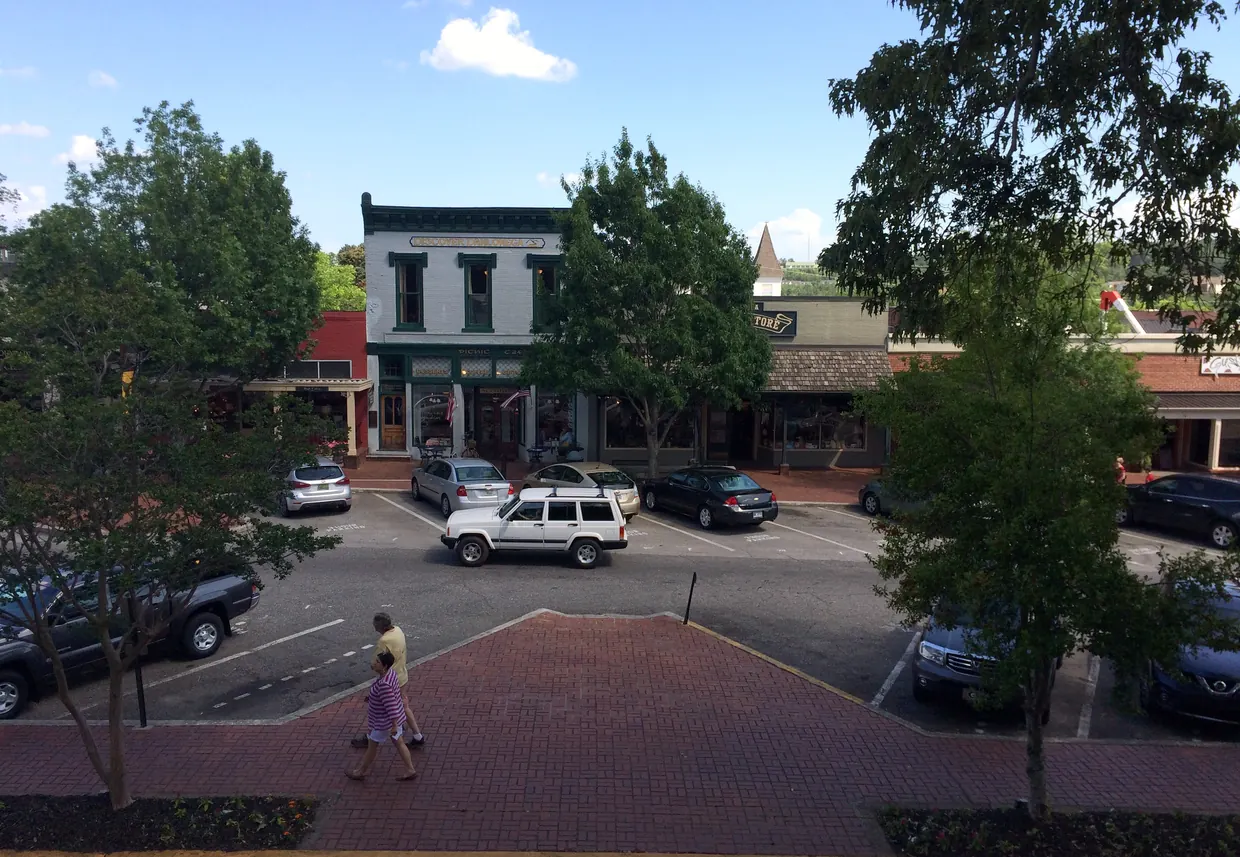 A row of two-storey shopfronts with awnings across an angle-parked lot, seen from an upper window, with two people crossing a brick plaza between young trees on a sunny day.